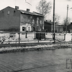na pierwszym planie brukowana ulica z torami tramwajowymi, w głębi jednopiętrowy dom mieszkalny ze spadzistym dachem, za nim parterowy budynek mieszkalny, wzdłuż ulicy odgarnięty zabrudzony śnieg