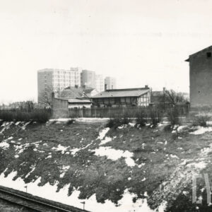 nasyp kolejowy, poniżej fragment torów kolejowych, na nasypie z prawej fragment budynku mieszkalnego, w środku zabudowania gospodarcze, w głębi wysokie bloki mieszkalne