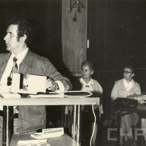 mężczyzna stojący przy rzutniku, za nim, w głębi siedzą trzy kobiety