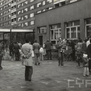 grupa ludzi stojąca przed niskim budynkiem, w głębi blok mieszkalny