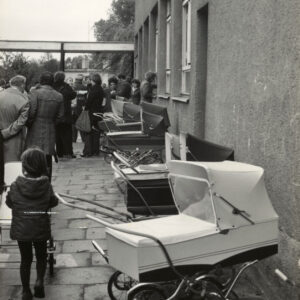 grupa ludzi stojąca przed niskim budynkiem, pośrodku wystawione dziecięce wózki