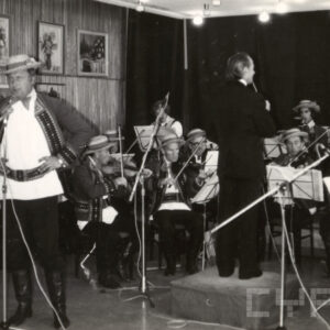 grupa muzyków w strojach ludowych gra na instrumentach ludowych, dyryguje mężczyzna w garniturze, z lewej solista w stroju ludowym śpiewa przy mikrofonie