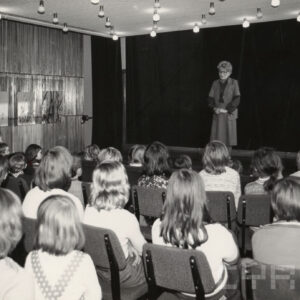 grupa dzieci siedzi na krzesłach w sali ze sceną, na scenie kobieta zwrócona przodem do dzieci