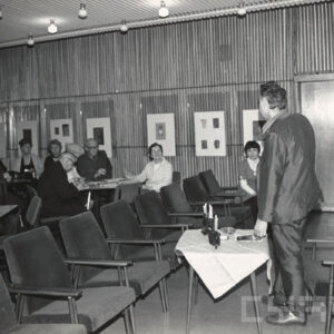 z prawej, tyłem stoi mężczyzna, przed nim niski stolik, nakryty jasnym obrusem, na nim palą się świece, w głębi grupy ludzi siedzą przy stolikach