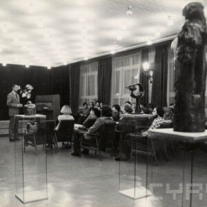 grupa ludzi siedzi i słucha zwróconego do nich mężczyzny, na pierwszym planie rzeźby na szklanych postumentach