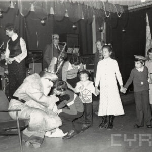 grupa dzieci, część z nich przebranych, trzyma się za ręce, z lewej przebrana osoba, przy niej pochylony chłopiec, w głębi scena z muzykami