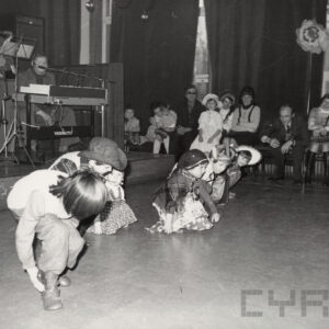 kilkoro dzieci w pozycji przykucniętej, część z nich przebrana, za nimi na scenie muzycy, w głębi dzieci i dorośli siedzą na krzesłach