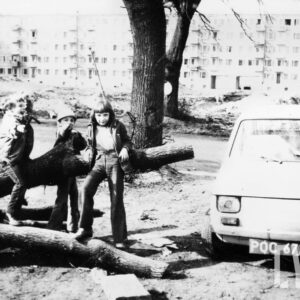 grupka dzieci bawiących się na powalonej gałęzi drzewa obok samochodu fiat 126p; w tle budowa bloku z wielkiej płyty