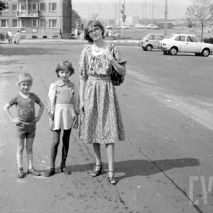 młoda kobieta z dwójką dzieci: synem i córką na parkingu samochodowym; w głębi samochody i budynek mieszkalny z wielkiej płyty 