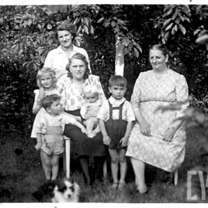 trzy dorosłe kobiety, trójka dzieci i niemowlę pozujący do fotografii na tle drzew, część z nich siedzi na ławce, część stoi, na dole fotografii widoczny pies