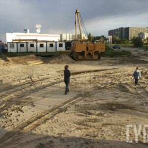 ludzie spacerujący po piaszczystym terenie, widoczna koparka i tymczasowe baraki dla robotników, w oddali budynki