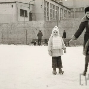 dziewczynka na łyżwach stoi obok mamy; w głębi budynek z wielkiej płyty