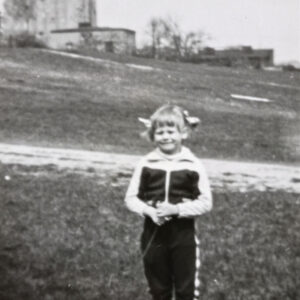 dziewczynka stojąca na trawie; w oddali siedmiokondygnacyjny budynek z betonu