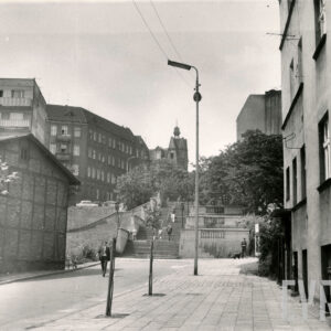 asfaltowa ulica z szerokim chodnikiem, w środku szerokie schody, idący po nich ludzie, dookoła nich kilkukondygnacyjne budynki