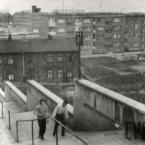 dwóch młodych mężczyzn wchodzi po schodach, za nimi dom drewniano-ceglany, w głębi czterokondygnacyjne bloki mieszkalne