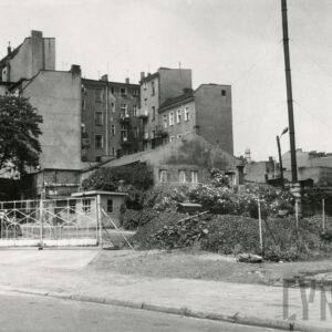 na pierwszym planie ogrodzony teren, pośrodku niski dom ze skośnym dachem, w głębi kilkukondygnacyjne kamienice