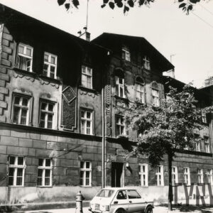 dwupiętrowa kamienica z mieszkalnym poddaszem, przed budynkiem drzewa, na chodniku zaparkowany samochód osobowy