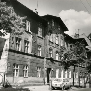 dwupiętrowa kamienica z mieszkalnym poddaszem, przed budynkiem drzewa, na chodniku zaparkowane samochody osobowe