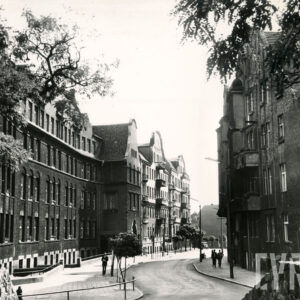 asfaltowa ulica, po jej stronach kilkukondygnacyjne budynki, po chodniku spacerują ludzie