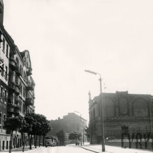 asfaltowa ulica, z lewej kilkukondygnacyjne kamienice, przed nimi drzewa, z prawej, w oddali dwukondygnacyjny zdobiony budynek mieszkalny w formie willi miejskiej