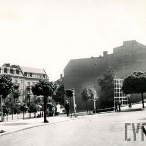 asfaltowa ulica, z jej lewej strony pusty plac, w głębi ściana bez okien kilkukondygnacyjnego budynku