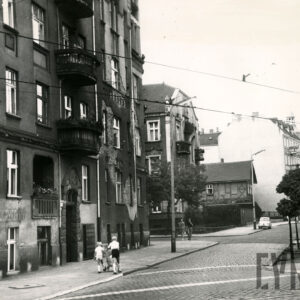 brukowana ulica, przy niej kilkukondygnacyjna kamienica, po chodniku spacerują ludzie