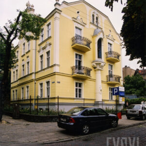 dwupiętrowa willa z suteryną i strychem bogato zdobioną dekoracją rzeźbiarską, stoi w małym ogrodzie otoczonym metalowym płotem