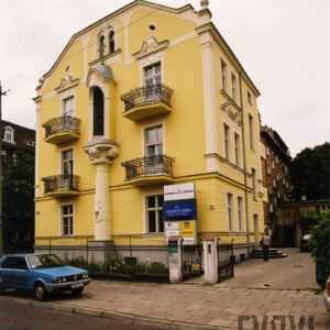 dwupiętrowa willa z suteryną i strychem bogato zdobioną dekoracją rzeźbiarską, pośrodku kolumna na niej w niszy figura świętego, dom stoi w małym ogrodzie otoczonym metalowym płotem