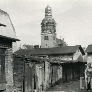 w jednym rzędzie stoją niskie drewniane szopy, z prawej idzie kobieta, w głębi wieża kościoła, w jej górnej partii rusztowania