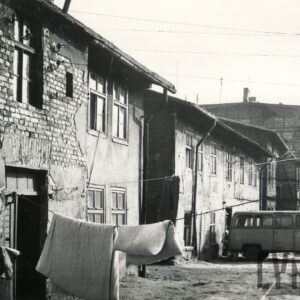 podwórze przy dwukondygnacyjnych budynkach murowanych, z odpadającym z elewacji tynkiem, na rozwieszonych sznurach wywieszona pościel, w głębi mały samochód dostawczy