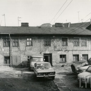 zaniedbany dwukondygnacyjny budynek ze spadzistym dachem krytym papą, przed budynkiem zaparkowane samochody