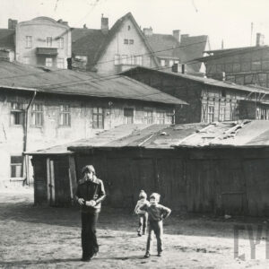 na pierwszym palnie w jednym rzędzie niskie drewniane szopy, przed nimi trójka dzieci, starszy chłopiec i dwoje młodszych, w głębi dwukondygnacyjne zaniedbane domy mieszkalne