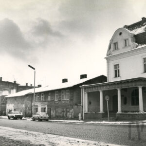na pierwszym planie brukowana ulica, po niej jadą lub parkują przy niej samochody, wzdłuż ulicy dwukondygnacyjne proste domy, z prawej piętrowy budynek z mansardowym dachem i gankiem z kolumnami, zimowa sceneria