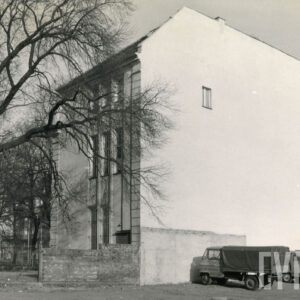 trzykondygnacyjny budynek z widocznymi dwiema ścianami, na jednej długie, wąskie okna, na drugiej pusta ściana tylko z jednym małym oknem, przy niej zaparkowane dwa samochody dostawcze, z lewej duże drzewo pozbawione liści
