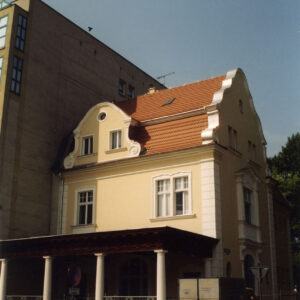 piętrowy budynek z mansardowym dachem z wejściem w formie ganku z kolumnami, które wspierają daszek, z lewej fragment wysokiej prostej ściany