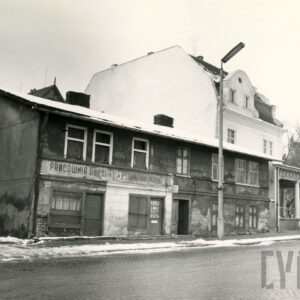 zniszczony dwukondygnacyjny, prosty, długi budynek, za nim piętrowy budynek z mansardowym dachem z wejściem w formie ganku z kolumnami, które wspierają daszek, zimowa sceneria