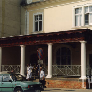 fragment piętrowego budynku z mansardowym dachem z wejściem w formie ganku z kolumnami, które wspierają daszek, przed budynkiem samochody i wychodzący ludzie