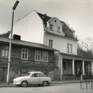 zaniedbany, dwukondygnacyjny prosty budynek, stojący przy brukowanej ulicy, obok niego zadbany, jasny wyższy budynek z mansardowym dachem i gankiem z kolumnami