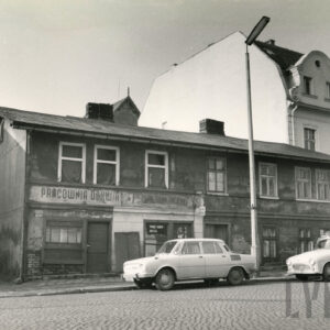 zaniedbany, dwukondygnacyjny prosty budynek, stojący przy brukowanej ulicy, za nim zadbany, wyższy, jasny budynek