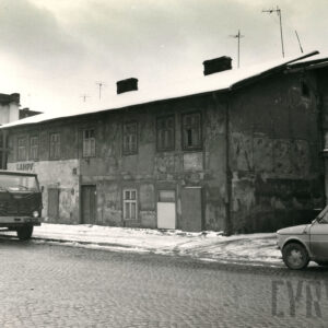 zaniedbany, dwukondygnacyjny prosty budynek, stojący przy brukowanej ulicy, wzdłuż niej zaparkowane dwa samochody, jeden z nich ciężarowy