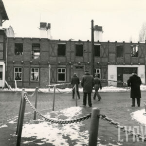 dwukondygnacyjny, drewniano-ceglany budynek pozbawiony dachu i częściowo ram okiennych, na pierwszym planie czterech mężczyzn przechodzących przez ulicę