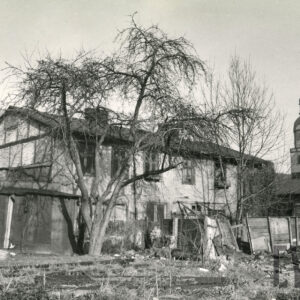 zaniedbane podwórze z dwukondygnacyjnym, prostym budynkiem, wokół małe szopy, przybudówki i walący się drewniany płot, na środku drzewo bez liści, z prawej fragment wieży kościoła