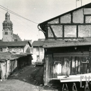 niskie drewniane szopy, z prawej fragment ściany budynku drewniano-ceglanego, w głębi, z lewej wieża kościoła