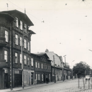 brukowana ulica, z lewej czterokondygnacyjny, narożny dom drewniano-ceglany, za nim dwukondygnacyjne proste domy mieszkalne