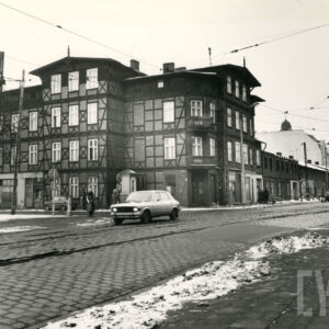 dwupiętrowy drewniano-ceglany dom narożny z mieszkalnym poddaszem, na pierwszym planie brukowana ulica z torami tramwajowymi i jadący nią samochód osobowy, resztki zalegającego śniegu