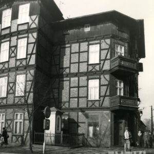 czterokondygnacyjny, drewniano-ceglany budynek z drewnianym poddaszem, w narożniku dwie kondygnacje z drewnianymi balkonami, w wyciętym narożniku budynku kapliczka ogrodzona niskim płotem, chodnikiem spacerują ludzie