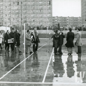 grupa mężczyzn na mokrym, betonowym korcie otoczonym przez dużą grupę dzieci, uczniów szkoły; w głębi budynki z wielkiej płyty