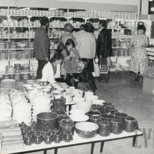 w głębi półki z ceramiką, przy nich ludzie, na pierwszym planie na niskich stolikach naczynia ceramiczne