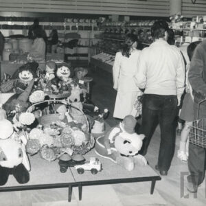 pośrodku niski stolik z zabawkami, za nim ludzie, z prawej mężczyzna z koszykiem, w głębi półki z towarami