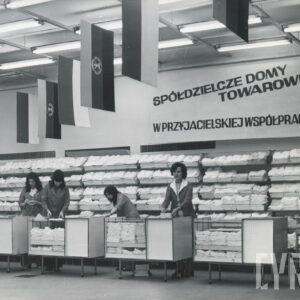 półki z artykułami odzieżowymi, przy ladach stoją sprzedawczynie, na ścianie duży napis „spółdzielcze domy towarowe w przyjacielskiej współpracy”